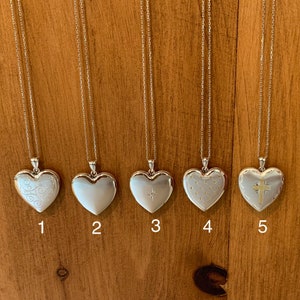 May include: Five silver heart-shaped locket necklaces with different engraved designs. The lockets are hanging from silver chains against a wooden background.
