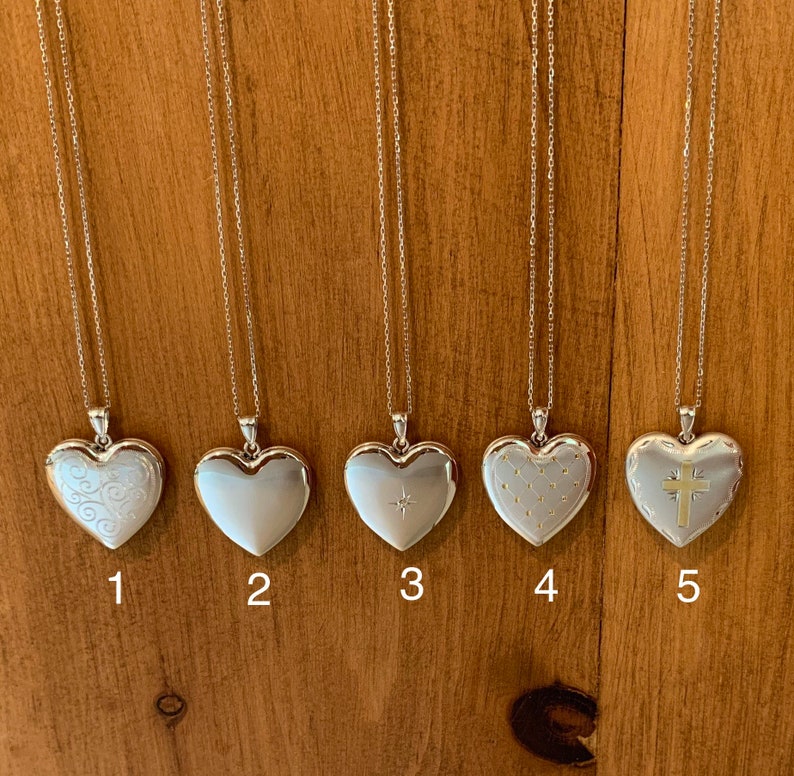 May include: Five silver heart-shaped locket necklaces with different engraved designs. The lockets are hanging from silver chains against a wooden background.