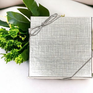 May include: A silver gift box with a silver ribbon tied in a bow. The box is sitting on a white surface with green leaves.