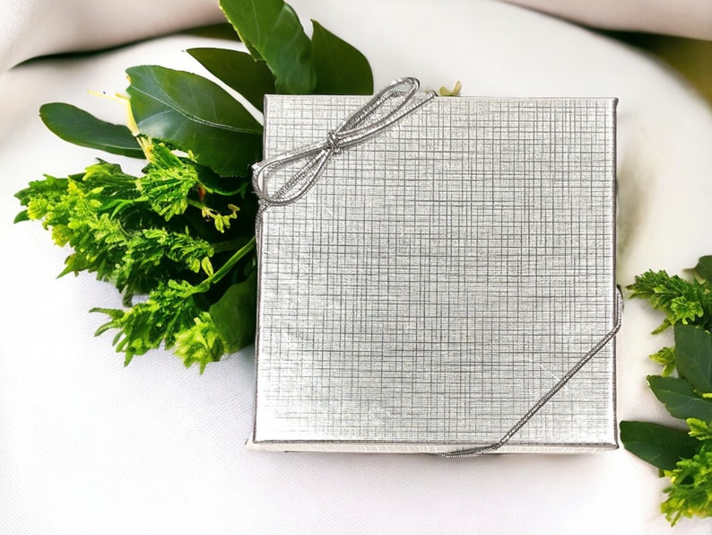 May include: A silver gift box with a silver ribbon tied in a bow. The box is sitting on a white surface with green leaves.
