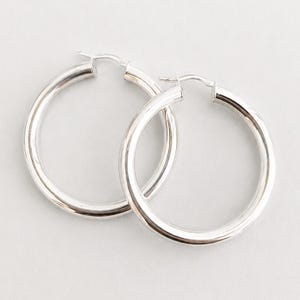 May include: A pair of polished silver hoop earrings with a smooth, rounded tubular design. Each earring features a secure hinged post closure at the top, creating a continuous circular silhouette with a bright, reflective metallic finish.