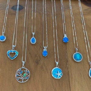 May include: Ten silver necklaces with blue opal pendants. The pendants are in various shapes, including a heart, a tree, a starfish, a teardrop, and a circle. The necklaces are all displayed on a wooden surface.