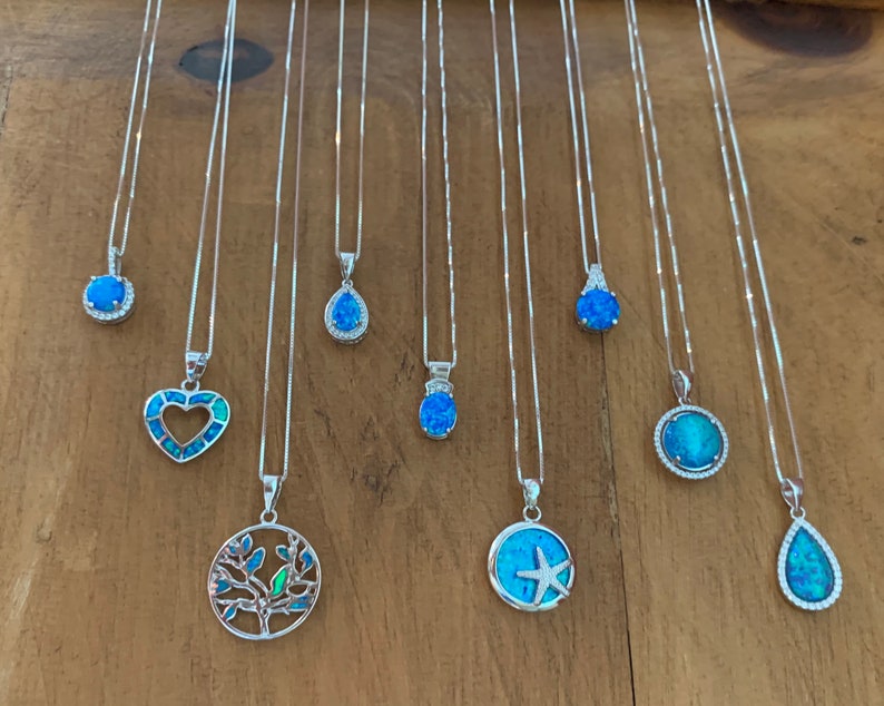 May include: Ten silver necklaces with blue opal pendants. The pendants are in various shapes, including a heart, a tree, a starfish, a teardrop, and a circle. The necklaces are all displayed on a wooden surface.