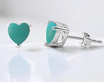 Puede incluir: Pendientes de botón con piedras turquesas en forma de corazón engastadas en metal plateado. Los pendientes se muestran sobre una superficie blanca, resaltando su diseño.