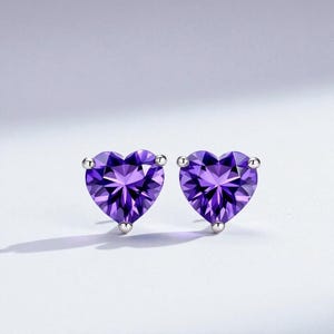 May include: A pair of stud earrings featuring heart-shaped, faceted purple gemstones. The earrings have a silver-colored post and backing. The gemstones are a deep violet color and are set against a white background.