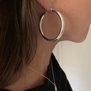 May include: A large, polished silver hoop earring with a smooth, rounded tubular design. The earring features a classic circular shape and a reflective metallic finish, worn through the earlobe. The piece is simple, circular, and silver-toned.