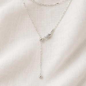 May include: A silver-toned necklace featuring a delicate chain with elongated rectangular links. The design includes an adjustable sliding mechanism with a small, round bead at the end of a dangling chain segment, finished with a secure lobster clasp.