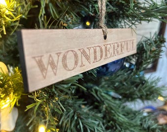 May include: A wooden Christmas ornament with the word "WONDERFUL" engraved in a decorative font. The ornament is rectangular and hangs from a twine string, set against a backdrop of a Christmas tree with lights.
