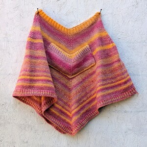 May include: A hand-knitted poncho featuring a vibrant gradient of orange, magenta, and purple horizontal stripes. The garment has a V-shaped neckline, a central V-shaped pocket on the front, and ribbed edges along the collar and hemline.