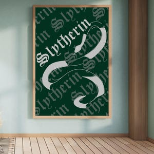 May include: A framed poster with a green background featuring the word "Slytherin" in white, stylized lettering. A white serpent design is intertwined with the text. The poster is in a wooden frame and hangs on a light blue wall.