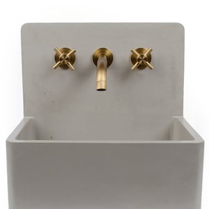 Puede incluir: Un lavabo de hormig&oacute;n con un grifo simple y dos botones. El lavabo es de color gris claro y el grifo y los botones son de color lat&oacute;n.