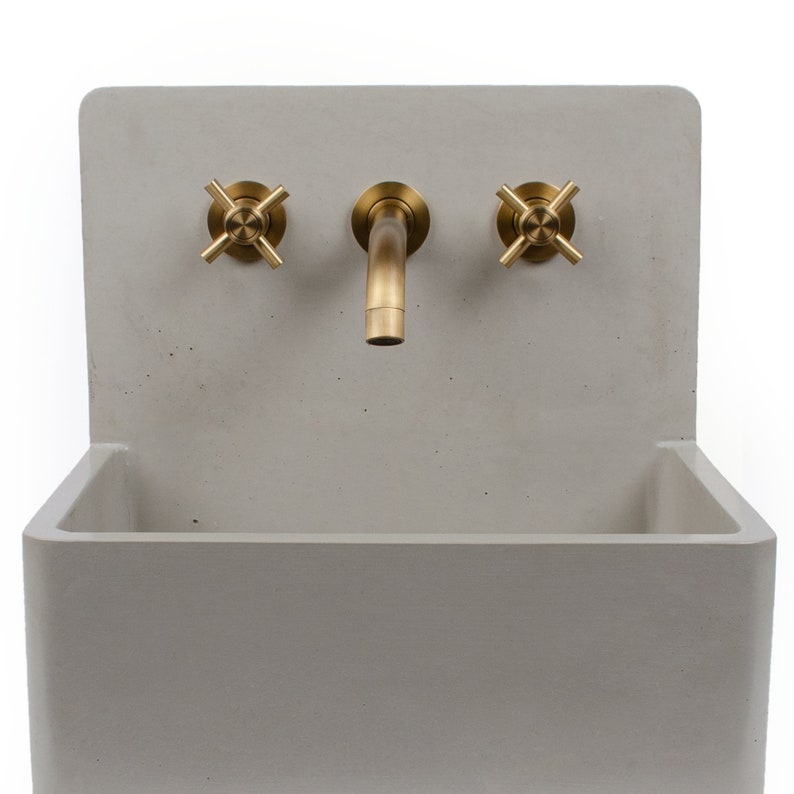 Puede incluir: Un lavabo de hormig&oacute;n con un grifo simple y dos botones. El lavabo es de color gris claro y el grifo y los botones son de color lat&oacute;n.