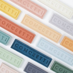 May include: A collection of colorful rectangular tiles with the word "CONCETTI" written on them in a white font. The tiles are arranged in a staggered pattern on a white background.