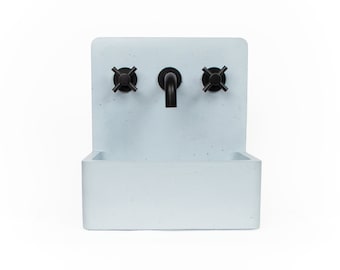 Puede incluir: Un lavabo de pared de hormigón blanco con un grifo negro y dos manijas negras en forma de cruz.