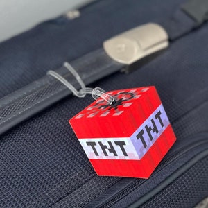 May include: Red luggage tag with a Minecraft TNT block design. The tag has the word "TNT" written on it twice.