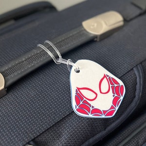 May include: A white luggage tag with a red and blue spider-themed design. The tag is attached to a dark blue suitcase with a clear plastic loop. The design features a spider mask.