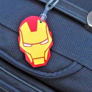 Puede incluir: Una etiqueta de equipaje roja y amarilla con una imagen de la cara de Iron Man. La etiqueta est&aacute; unida a una maleta azul.