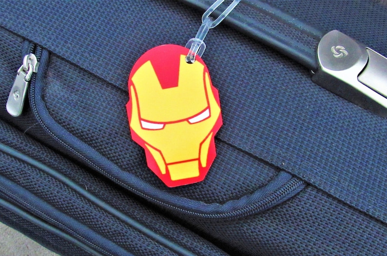 Puede incluir: Una etiqueta de equipaje roja y amarilla con una imagen de la cara de Iron Man. La etiqueta est&aacute; unida a una maleta azul.