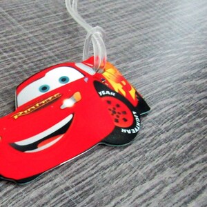 May include: Red cartoon car luggage tag with a smiling face and blue eyes. The car has the words "Lightning McQueen" on the side and "YEAR" and "LIGHTNING" on the tire.