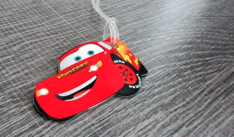 May include: Red cartoon car luggage tag with a smiling face and blue eyes. The car has the words "Lightning McQueen" on the side and "YEAR" and "LIGHTNING" on the tire.
