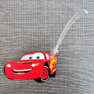 May include: Red cartoon race car luggage tag with the text "Lightning McQueen" and "Year" on the side. The tag is attached to a clear plastic loop.