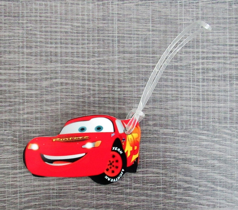 May include: Red cartoon race car luggage tag with the text "Lightning McQueen" and "Year" on the side. The tag is attached to a clear plastic loop.