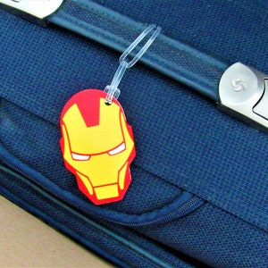 Puede incluir: Una maleta azul con una cremallera y una correa negra. Una etiqueta de equipaje roja y amarilla con una cara de superh&eacute;roe est&aacute; unida a la correa.