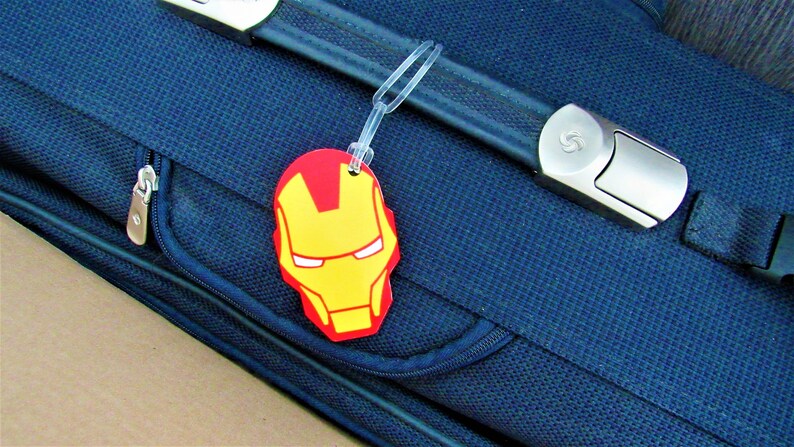 Puede incluir: Una maleta azul con una cremallera y una correa negra. Una etiqueta de equipaje roja y amarilla con una cara de superh&eacute;roe est&aacute; unida a la correa.