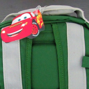 May include: Green backpack with gray straps and a red Lightning McQueen character tag attached to the top. The tag has the words "Team Lightning McQueen" and "Goodyear" on it.