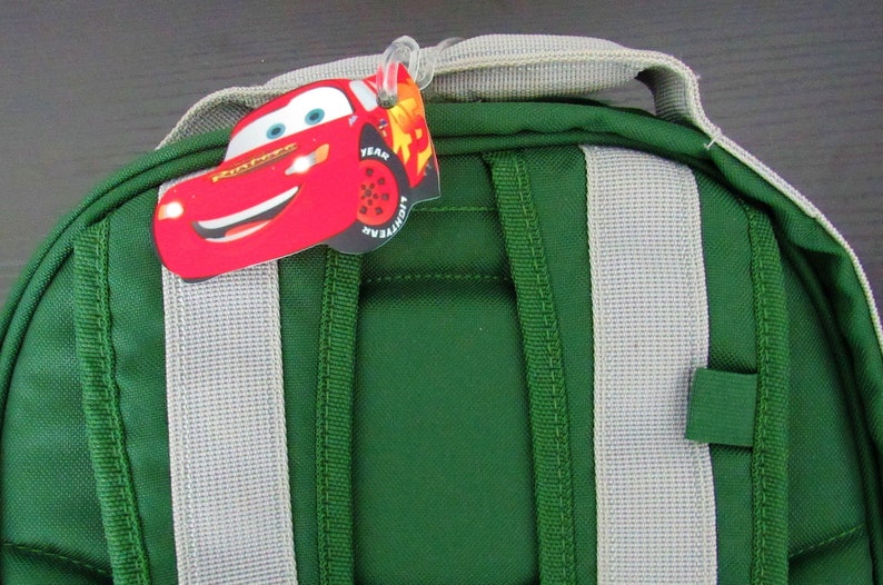 May include: Green backpack with gray straps and a red Lightning McQueen character tag attached to the top. The tag has the words "Team Lightning McQueen" and "Goodyear" on it.
