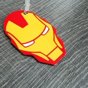 Puede incluir: Una etiqueta de equipaje roja y amarilla con un rostro estilizado de Iron Man, un superh&eacute;roe del universo Marvel.
