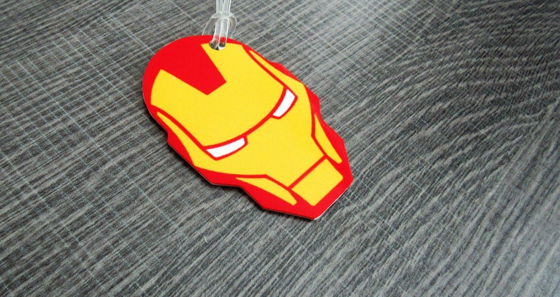 Puede incluir: Una etiqueta de equipaje roja y amarilla con un rostro estilizado de Iron Man, un superh&eacute;roe del universo Marvel.