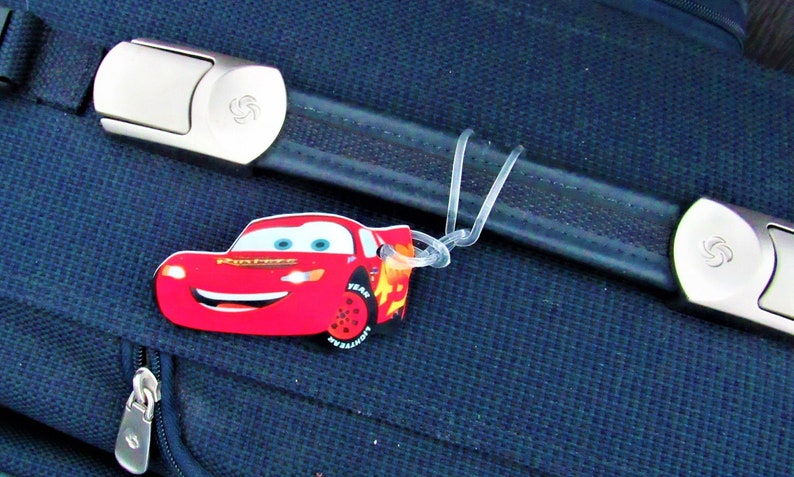 May include: A red cartoon car with a smiling face and the word "Lightning McQueen" on the side is attached to a blue suitcase with a silver handle.