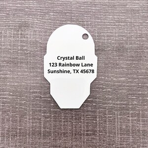 Puede incluir: Etiqueta blanca con bordes redondeados y un agujero en la parte superior. La etiqueta tiene el texto "Crystal Ball 123 Rainbow Lane Sunshine, TX 45678" impreso en ella.
