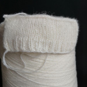 May include: A close-up of a white ball of yarn with a knitted fabric swatch on top. The yarn is wound tightly and the fabric is soft and fluffy.