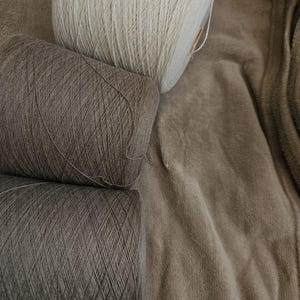 May include: Three yarn skeins in beige and brown tones are arranged on a soft, textured surface. The top skein is a light beige, with the other two in shades of brown. The tightly wound yarn suggests it's suitable for knitting or crochet projects.