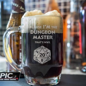 Peut inclure: Une chope à bière en verre avec un liquide noir et de la mousse blanche. La chope a une poignée et est gravée avec le texte "Because I'm the Dungeon Master That's Why." et un dé à 20 faces.