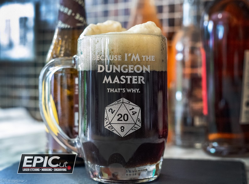 May include: A glass beer mug with a black liquid and white foam. The mug has a handle and is engraved with the text "Because I'm the Dungeon Master That's Why." and a 20-sided die.