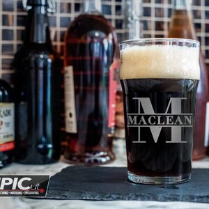 May include: A personalised pint glass featuring a large etched letter "M" with the name "Maclean" underneath. The glass is filled with dark beer and rests on a black slate coaster.
