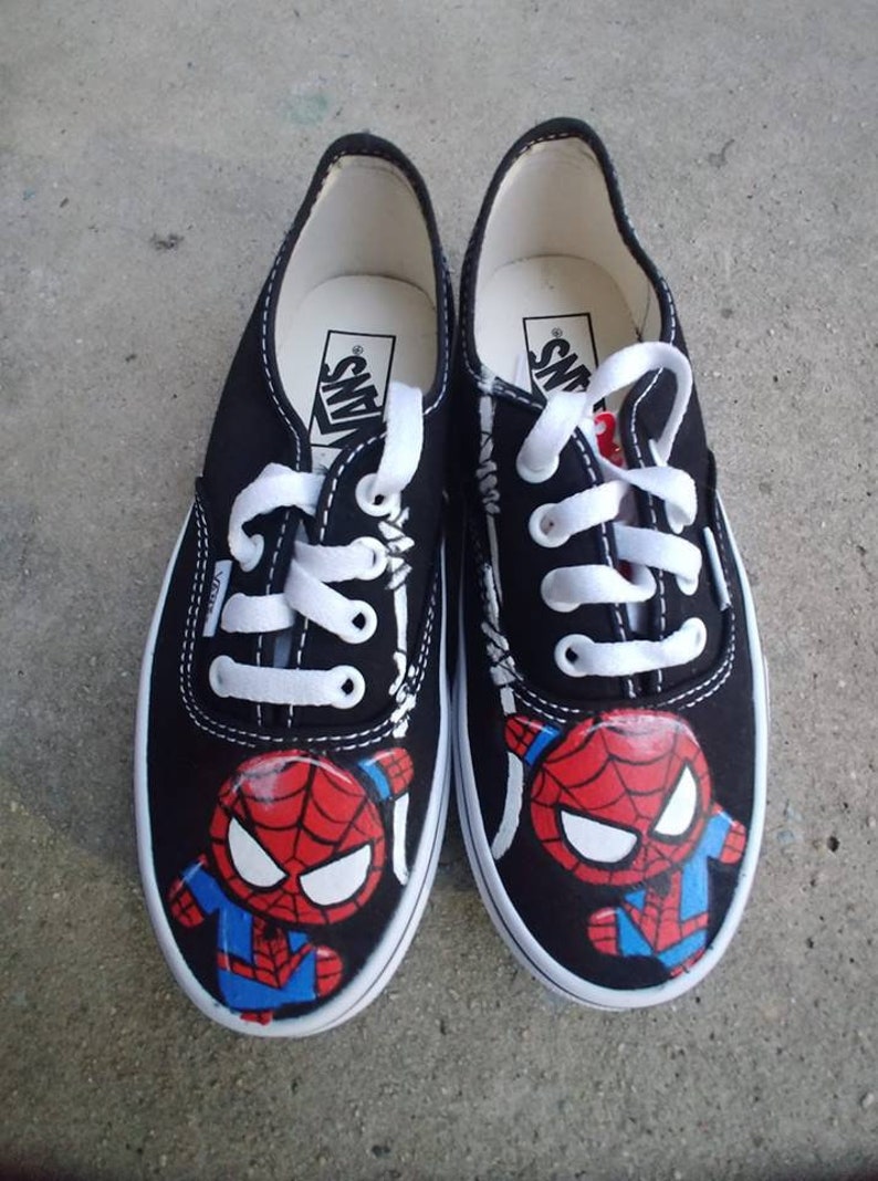 May include: Black canvas Vans sneakers with a hand-painted cartoon Spider-Man design. The shoes have white laces and white stitching. The Spider-Man design is in red, blue, and black.