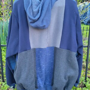 May include: A blue patchwork hoodie with a hood, hanging on a black hanger. The hoodie features panels of various shades of blue and grey. The garment is made of a soft, comfortable material.
