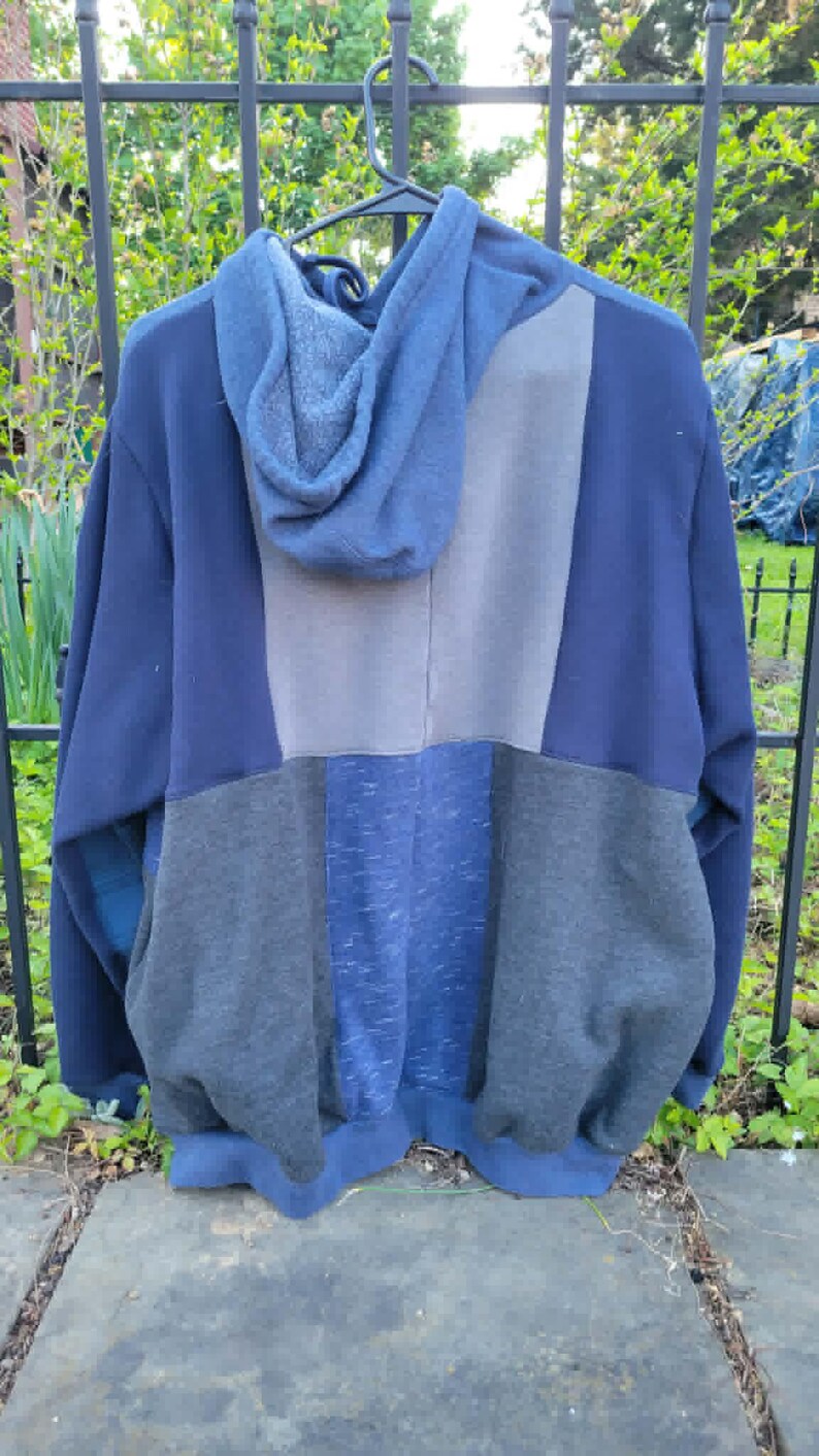 May include: A blue patchwork hoodie with a hood, hanging on a black hanger. The hoodie features panels of various shades of blue and grey. The garment is made of a soft, comfortable material.