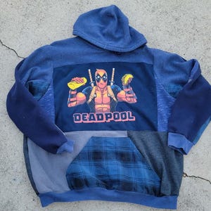 May include: A blue hooded sweatshirt with a Deadpool graphic. The graphic depicts Deadpool holding tacos and swords, with the word "DEADPOOL" in pink. The sweatshirt has a patchwork design with blue and grey panels.