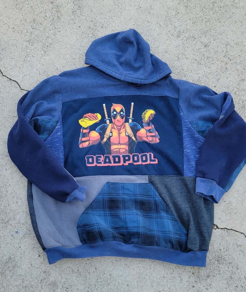 May include: A blue hooded sweatshirt with a Deadpool graphic. The graphic depicts Deadpool holding tacos and swords, with the word "DEADPOOL" in pink. The sweatshirt has a patchwork design with blue and grey panels.