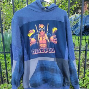 May include: A blue patchwork hoodie with a Deadpool graphic and the text "DEADPOOL". The hoodie has a hood, a front pocket, and a mix of blue, grey, and plaid fabric panels. The Deadpool graphic shows the character holding tacos.