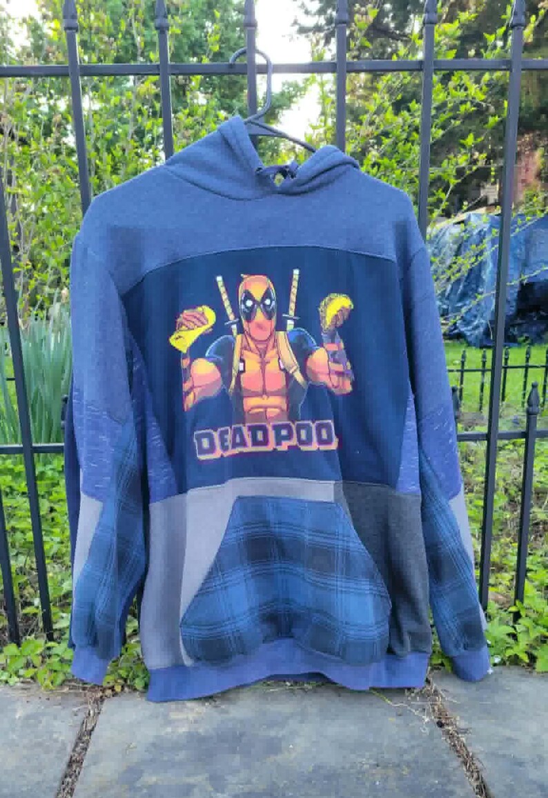 May include: A blue patchwork hoodie with a Deadpool graphic and the text "DEADPOOL". The hoodie has a hood, a front pocket, and a mix of blue, grey, and plaid fabric panels. The Deadpool graphic shows the character holding tacos.