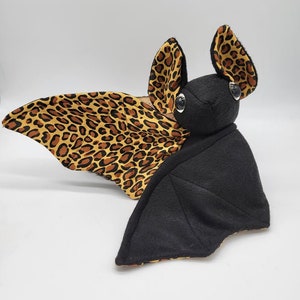 May include: A black plush bat with brown and yellow leopard print wings. The bat has large, black eyes and a small, black nose.