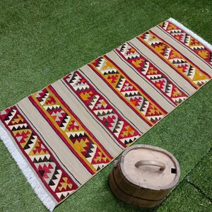 May include: A beige rug with a geometric pattern in shades of red, yellow, and black. The rug has a fringe border and is laid out on a green grassy surface.
