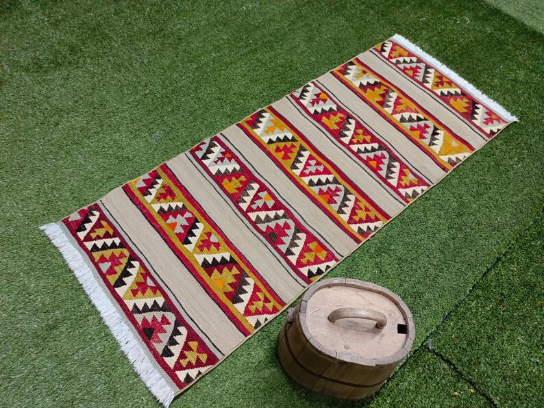 May include: A beige rug with a geometric pattern in shades of red, yellow, and black. The rug has a fringe border and is laid out on a green grassy surface.