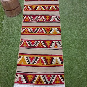 May include: A long, rectangular rug with a white fringe. The rug has a geometric pattern in shades of red, yellow, black, and gray.